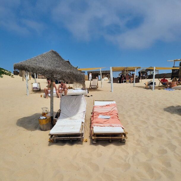 Two casual sun beds and a glass pitcher of red wine sangria under a palm straw umbrella on a Lisbon beach