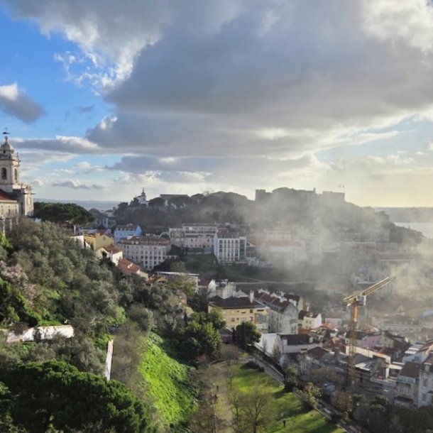 Dramatic sunset view from Miradouro da Senhora do Monte showing smoke rising from a building fire in Mouraria valley with Graça and Castelo hills framing the background above the Tejo River and Christ the King statue.