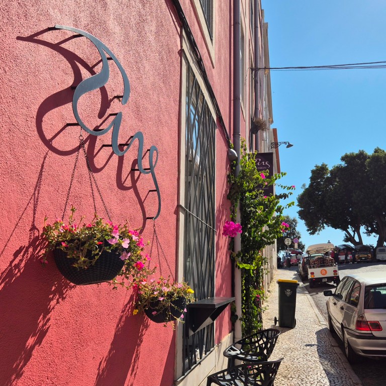 Vibrant pink bougainvillea and hanging petunias on the pink exterior facade beneath the Tings logo sign at our Graça boutique hotel Lisbon street front.